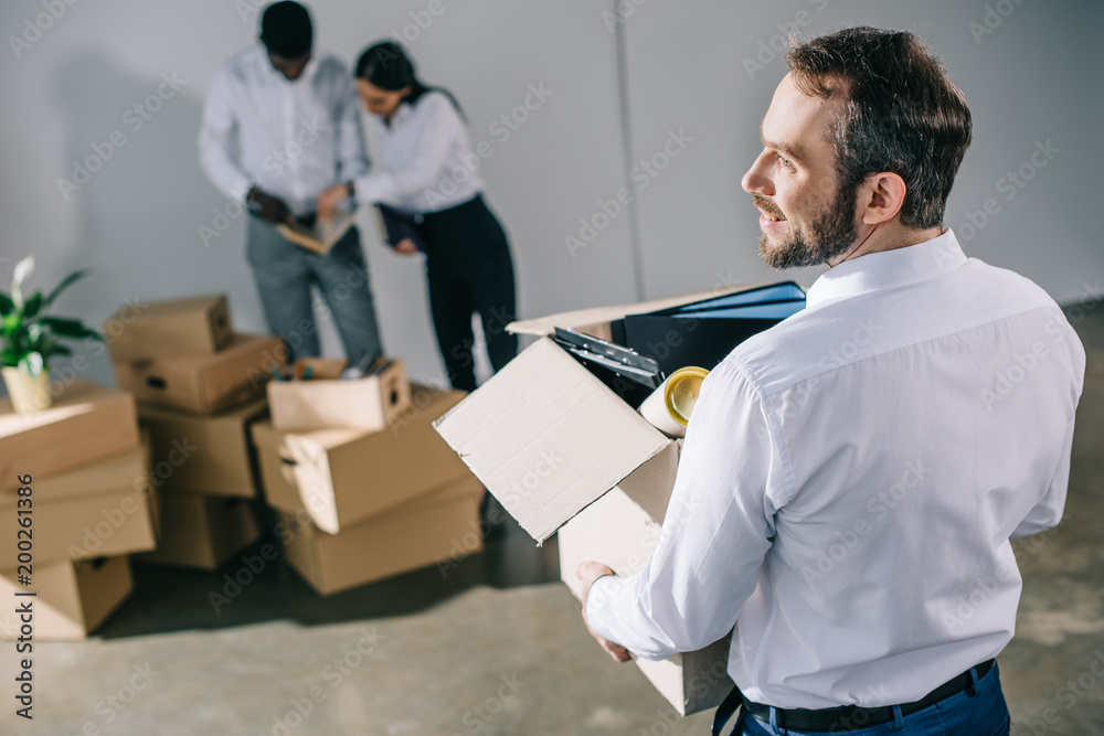 back view of businessman holding cardboard box with office supplies while colleagues unpacking boxes in new office back view of businessman holding cardboard box with office supplies while colleagues unpacking boxes in new office
