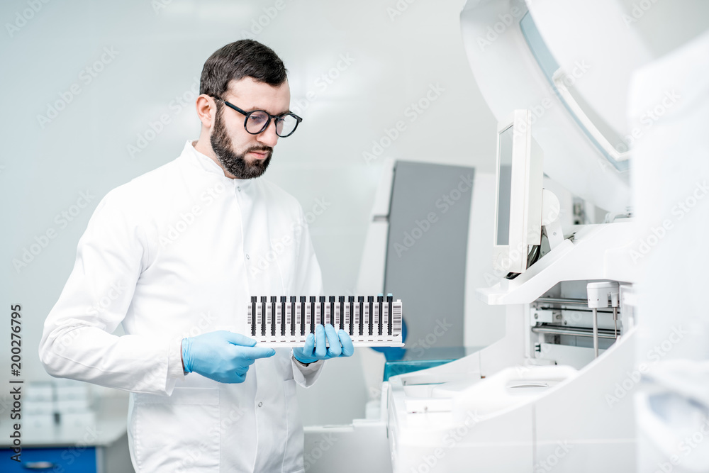 Male laboratory assistant in uniform holding test tubes near the analizer machine at the laboratory Male laboratory assistant in uniform holding test tubes near the analizer machine at the laboratory