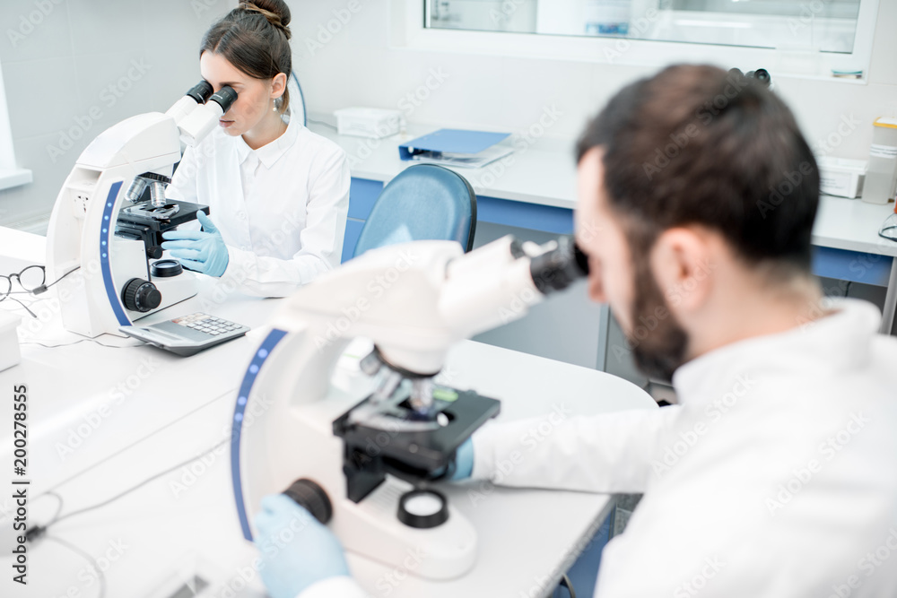 Two medics in uniform working with microscope making analysis at the laboratory office Two medics in uniform working with microscope making analysis at the laboratory office