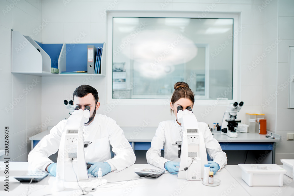 Naklejka premium Couple of medics in uniform working with microscope making analysis at the laboratory office