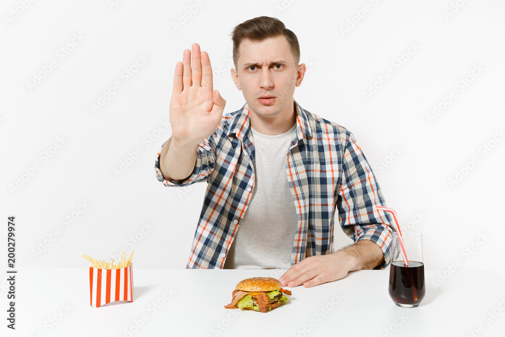 Severe man showing stop gesture with palm, sitting at table with burger, french fries, cola in glass isolated on white background. Proper nutrition or American classic fast food. Area with copy space. Severe man showing stop gesture with palm, sitting at table with burger, french fries, cola in glass isolated on white background. Proper nutrition or American classic fast food. Area with copy space.