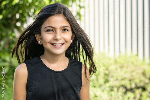 Portrait of a cute little girl in garden. Closeup portrait of a smiling brunette girl, blurred white fence and green plants in background.