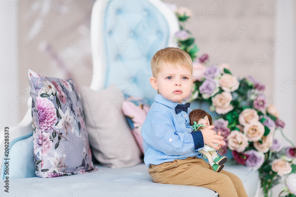 Portrait of a small cute boy gentleman in a blue shirt and with a tie. The child stands near a large window in a bright cozy house and holds a bouquet of flowers in his hand - a gift for the mother Portrait of a small cute boy gentleman in a blue shirt and with a tie. The child stands near a large window in a bright cozy house and holds a bouquet of flowers in his hand - a gift for the mother