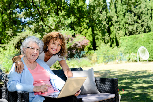 cheerful mature woman with mother elderly senior female sitting outdoor and playing with a laptop computer in retirement house hospital garden
