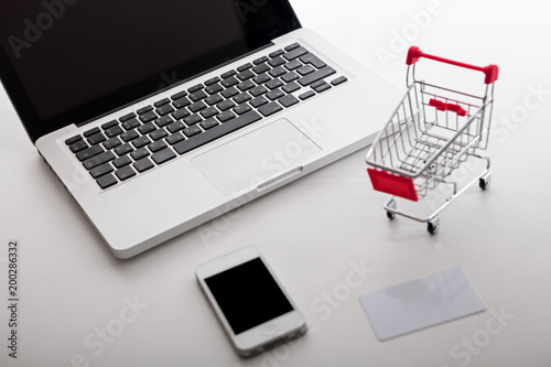Online shopping. Bank card nearby a laptop and mini shopping cart on white background top view.
