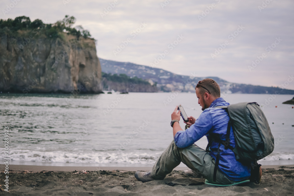 man, guy the photographer working look at the phone, receives a call, calls on the beach, backpack, concept of find information at travel to foregin country, communication, mountains on background