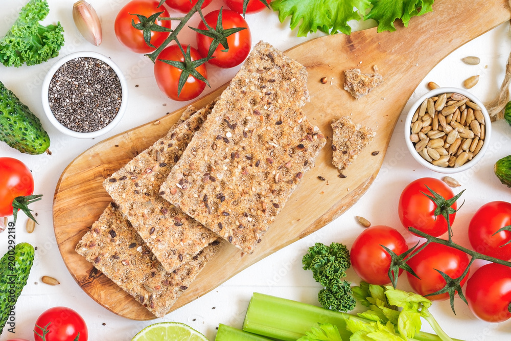 Top view of whole grain rye crispbreads on a wooden serving board surrounded fresh vegetables, greens and seeds over white background. Top view of whole grain rye crispbreads on a wooden serving board surrounded fresh vegetables, greens and seeds over white background.