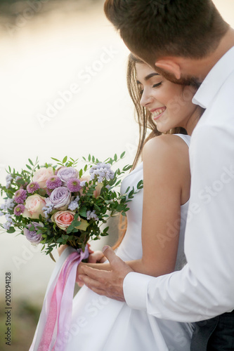 newly marriage beautiful couple, groom kissing his bride with wedding bouquet near a water at sunset. Close-up