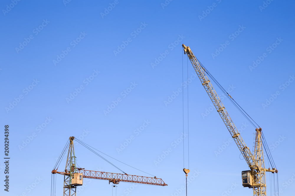 Front distant view of two cranes on a light blue sky background. One crane`s jib-arm is horizontal, the other one`s is placed diagonally with the lower sheave and hook hanging