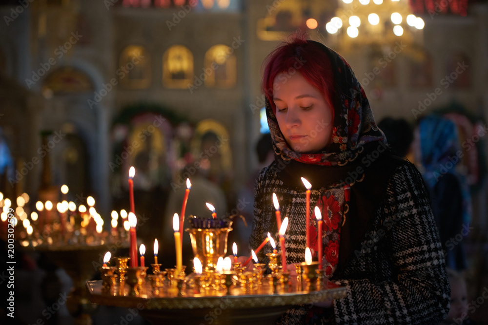 Praying young woman with candle near pedestal with many other candles in church by her hand to show her faith and esteem to God Praying young woman with candle near pedestal with many other candles in church by her hand to show her faith and esteem to God