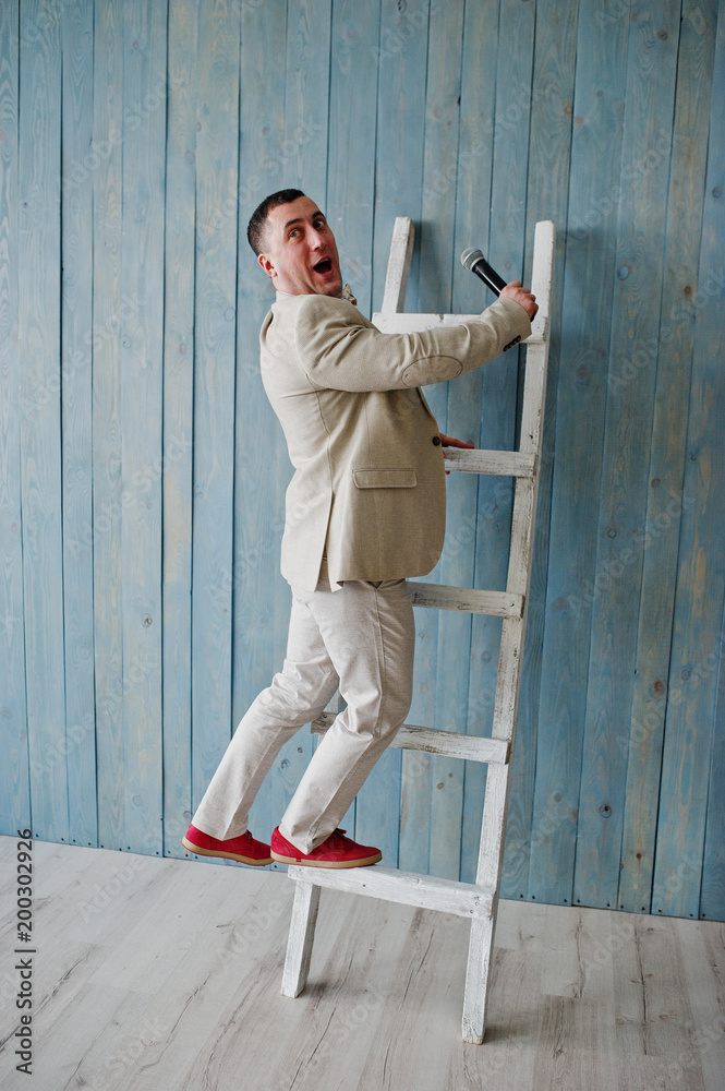 Handsome man in beige suit and pink shirt with microphone against ladder background on studio. Toastmaster and showman. Handsome man in beige suit and pink shirt with microphone against ladder background on studio. Toastmaster and showman.