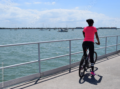 Fit African American woman riding bicycle on pier, water and sailboats in background.