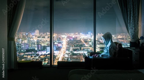 Hooded anonymous man working on laptop computer in dark hotel room, panoramic window with illuminated night city view in background. Timelapse, 4K UHD. Zoom in. photo