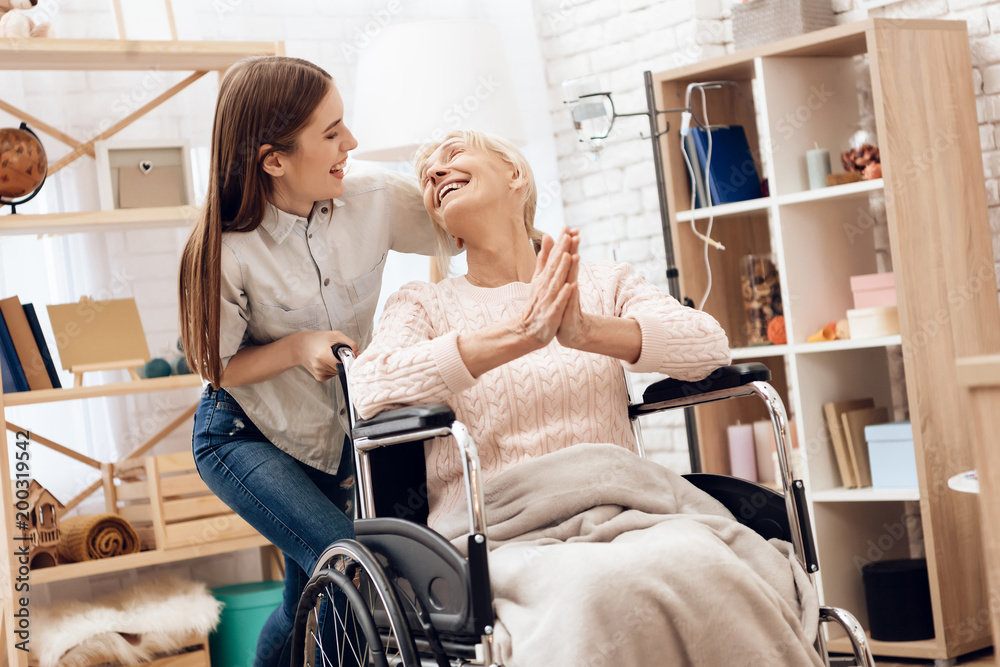Obraz premium Girl is nursing elderly woman at home. Girl is riding woman in wheelchair. Woman is enjoying herself.