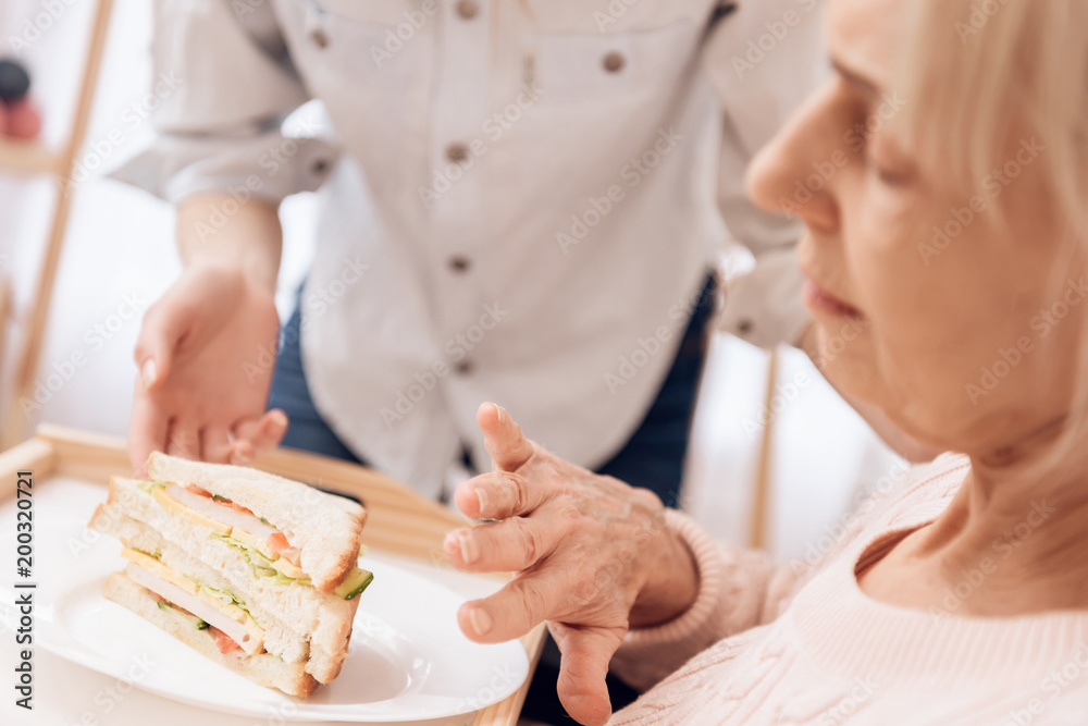 Girl is caring for elderly woman at home. Girl brings breakfast on tray. Woman is eating sandwitch. Girl is caring for elderly woman at home. Girl brings breakfast on tray. Woman is eating sandwitch.