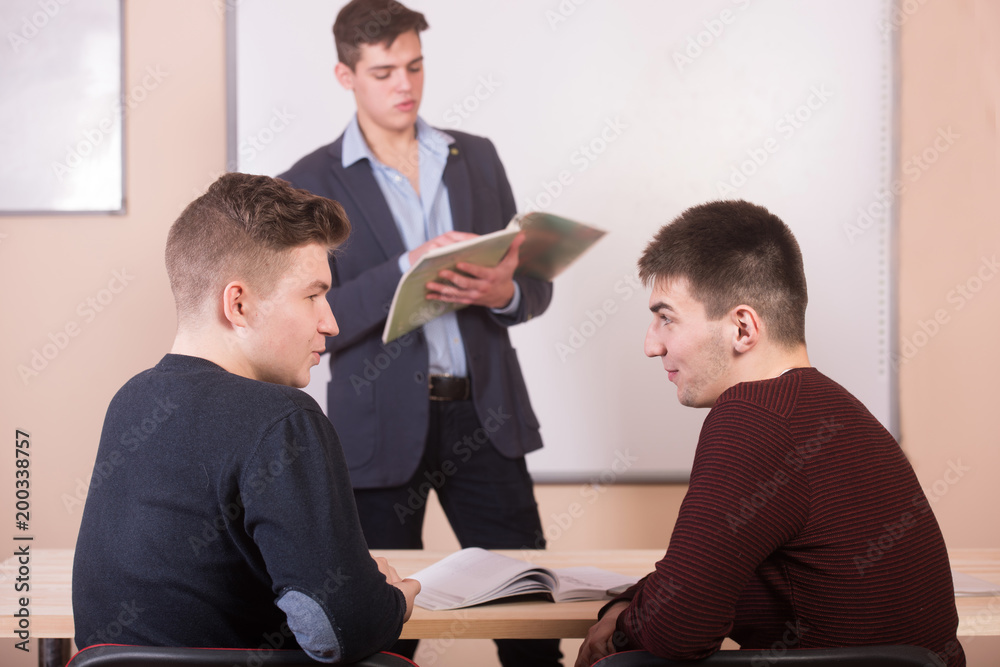 Discussion in the lesson. Youn teacher with book in background and two chatting students. Discussion in the lesson. Youn teacher with book in background and two chatting students.
