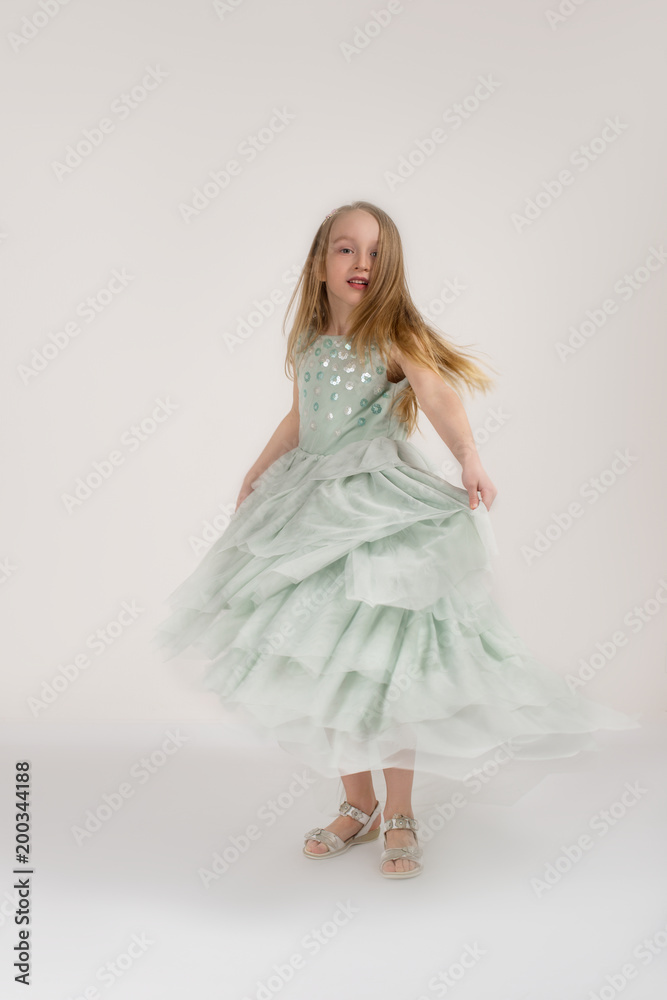 Naklejka premium Full length portrait of the dancing of the little girl of the blonde wearing green dress on a white background in studio.