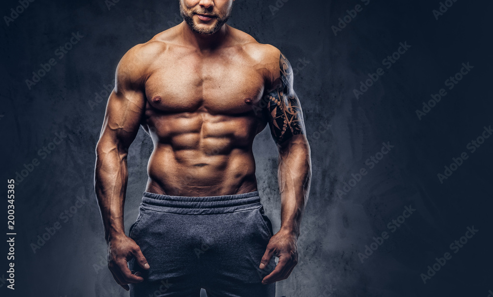 Fototapeta premium A handsome shirtless tattooed bodybuilder with stylish haircut and beard, wearing sports shorts, posing in a studio. Isolated on a dark background