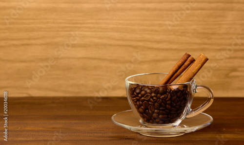 Fotografie A glass cup with coffee beans and cinamon stics on wooden background with copysp