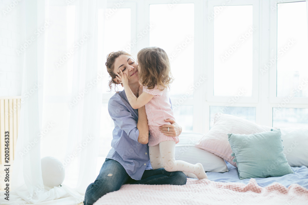 Young mother and her little daughter hugging and kissing on bed on a sunny morning. Soft pastel colors, pink and blue. Selective focus. Love and care. Happy family time on weekend. Mother's Day Young mother and her little daughter hugging and kissing on bed on a sunny morning. Soft pastel colors, pink and blue. Selective focus. Love and care. Happy family time on weekend. Mother's Day