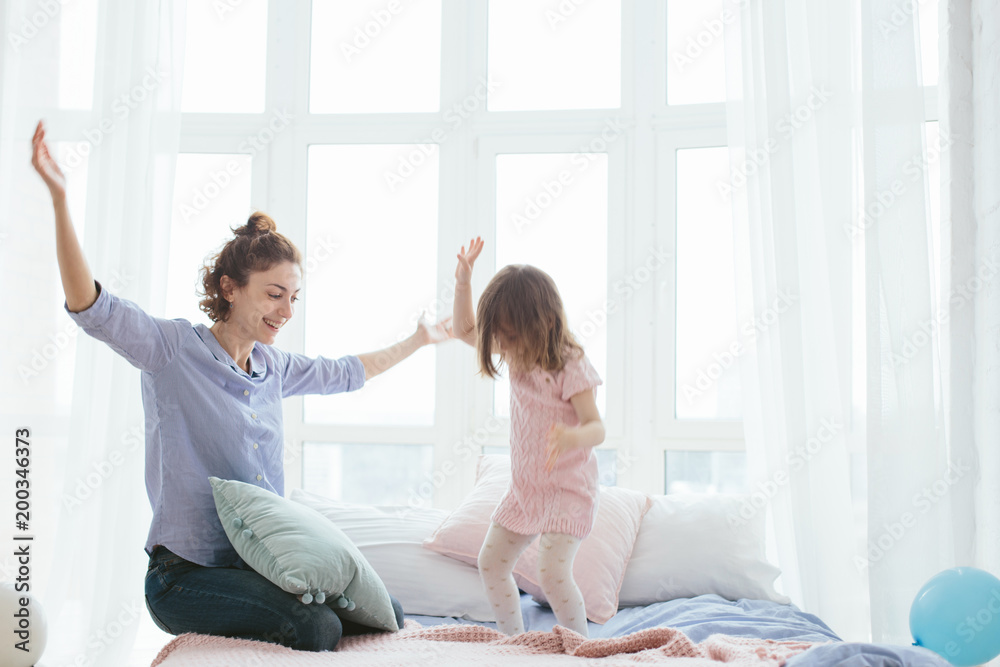 Young mother and her little daughter jumping on bed. Funny pillow fight. Soft pastel colors, pink and blue. Selective focus. Play together and enjoy the moment! Family time on weekend. Young mother and her little daughter jumping on bed. Funny pillow fight. Soft pastel colors, pink and blue. Selective focus. Play together and enjoy the moment! Family time on weekend.