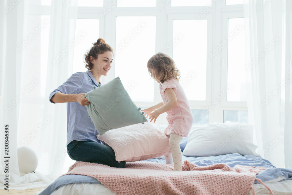 Young mother and her little daughter playing with cushions on bed. Funny pillow fight. Soft pastel colors, pink and blue. Selective focus. Play together and enjoy the moment! Family time on weekend. 