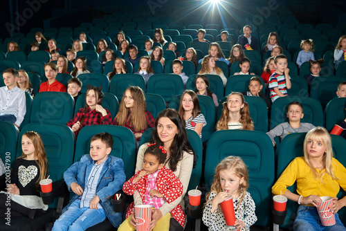 Photo of people watching movie in the cinema hall. They are very emotional, exited and satisfied. Some children eating popcorn or drinking fizzy drinks.