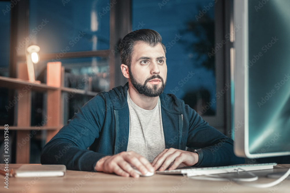 Accurate work. Serious meditative IT guy sitting at table while putting hand on mouse and gazing at screen Accurate work. Serious meditative IT guy sitting at table while putting hand on mouse and gazing at screen