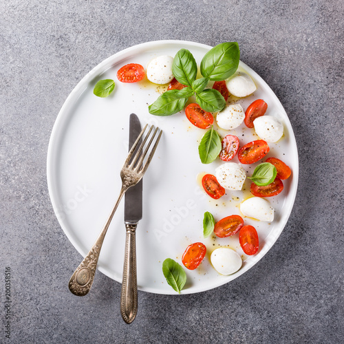 White plate of healthy classic delicious caprese salad with ripe tomatoes and mozzarella cheese with fresh basil leaves on gray concrete background with space for text. Italian food. Top view.