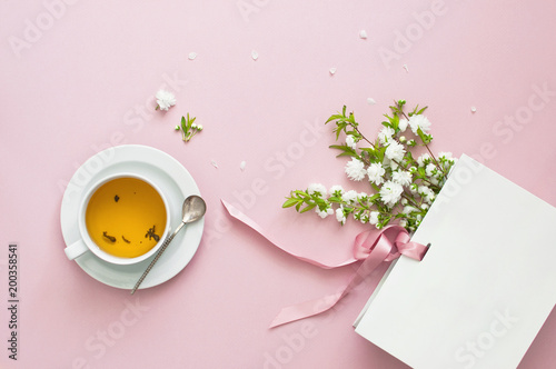 Flatlay cup of tea, white gift bag and spring flowers on a pink background. Beautiful breakfast. Workspace pastel colors. Greeting card with delicate flowers Pink floral background photo