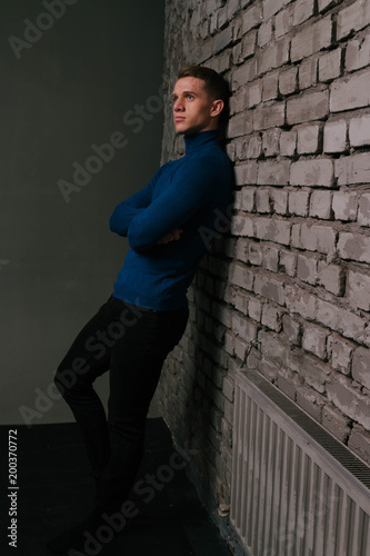 white attractive guy is nearby a brick wall background dressed in a blue sweater and black pants, next battery