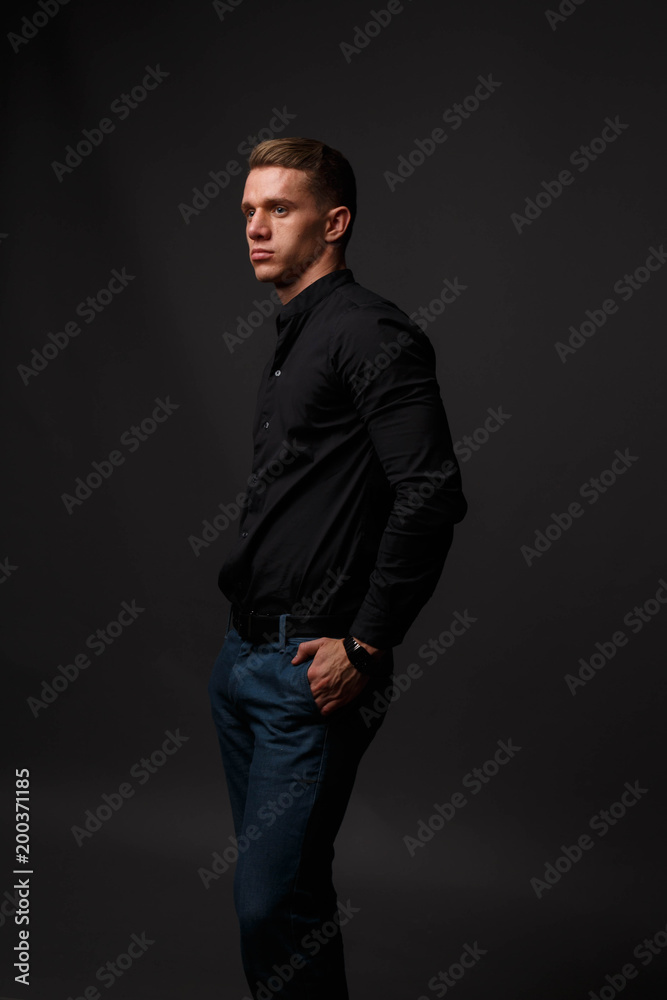 attractive white man in black shirt and blue pants stands on a dark background