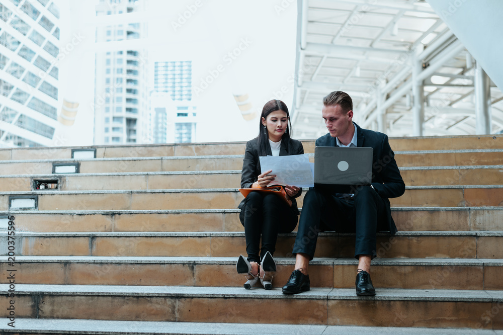 Asian business woman and Caucasian businessman sit on the staircase and hold a paper of sale report with laptop. concept of financial report and marketing plan for the future. Asian business woman and Caucasian businessman sit on the staircase and hold a paper of sale report with laptop. concept of financial report and marketing plan for the future.
