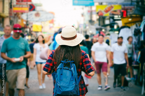 Young Asian woman with hat and backpack traveling in Khaosan Road among people, walking outdoor market in Bangkok, Thailand.