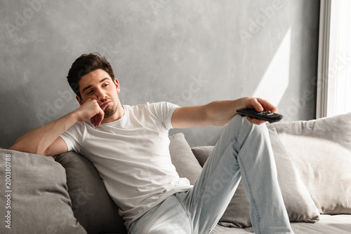 Photo of young bored man sitting on couch at home, and changing TV channels with disinterest