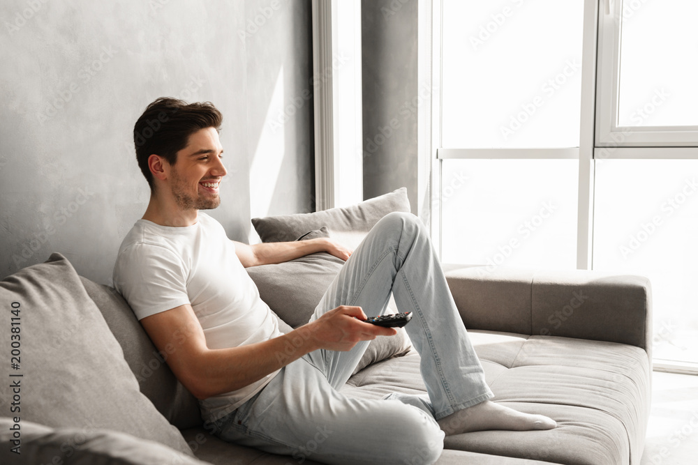 Image in profile of attractive brunette man sitting on sofa at home and watching television, with remote control in hand