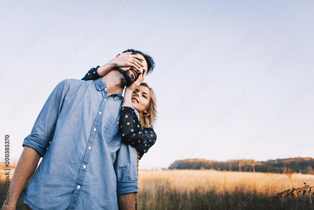 handsome guy with a beard in a denim shirt hugs and holds the hand of a beautiful girl in a blue dress and yellow scarf in a field at sunset. stylish couple sitting on a wooden fence and look around handsome guy with a beard in a denim shirt hugs and holds the hand of a beautiful girl in a blue dress and yellow scarf in a field at sunset. stylish couple sitting on a wooden fence and look around