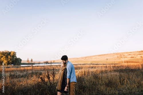 handsome guy with a beard in a denim shirt hugs and holds the hand of a beautiful girl in a blue dress and yellow scarf in a field at sunset. stylish couple sitting on a wooden fence and look around