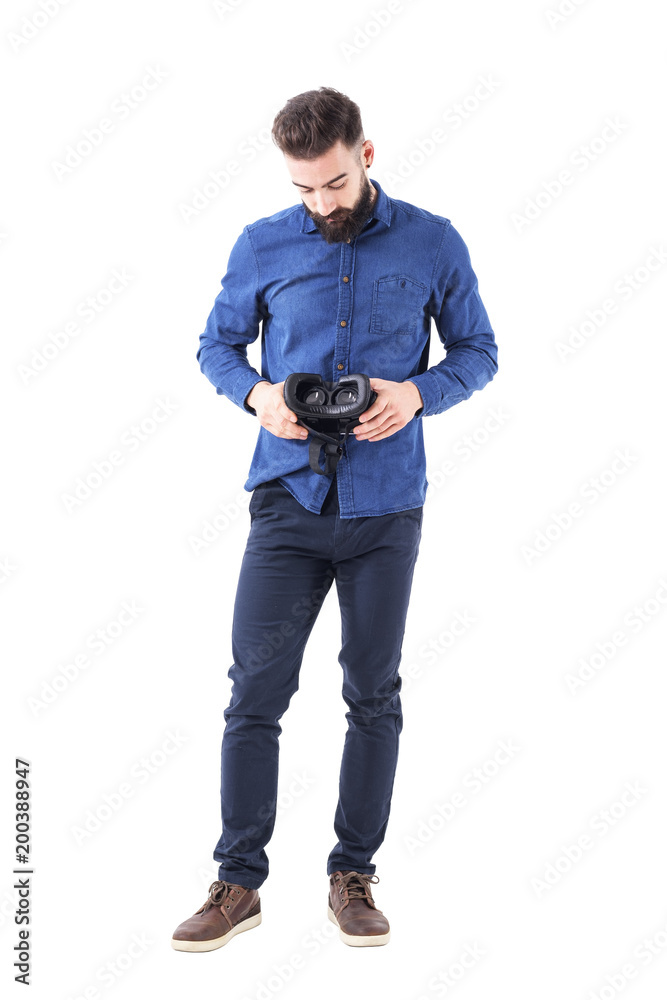 Fototapeta premium Front view of young bearded business man holding and looking down examining vr headset. Full body isolated on white background. 