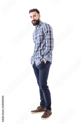 Happy young bearded man in checked shirt with hands in pockets smiling and looking at camera. Full body isolated on white background. 