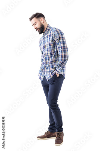 Cuadro en lienzo Shy handsome bearded young man in plaid shirt with hands in pockets smiling and looking down