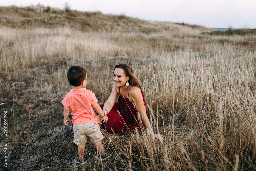 portret of a beautiful young mother in a long red dress with her little son walking on mountain sites on the sunset in summer, dry grass.Mom holds her son's hand and hugs