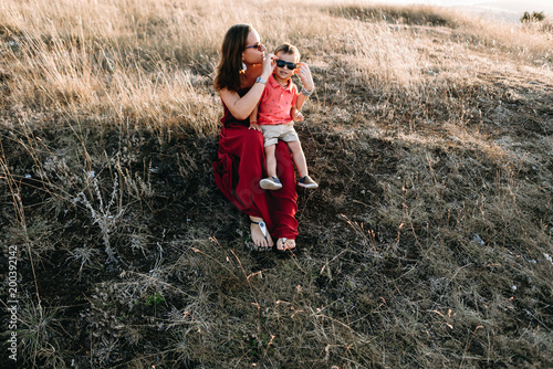 beautiful young mother in a long red dress with her little son walking on mountain sites on the sunset in summer, dry grass