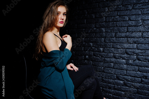 Handsome brunette girl wear on black and green jacket, sitting and posing on chair at studio against dark brick wall. Studio model portrait. © AS Photo Family