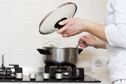 woman is cooking soup in kitchen. housewife prepares food at home. caucasian woman holds lid from saucepan in her hand and stirs soup in saucepan.