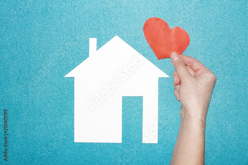 concept of love in family and at home. hand holds red heart over white paper house on blue background. Insurance of real estate. Loving and protecting the house.
