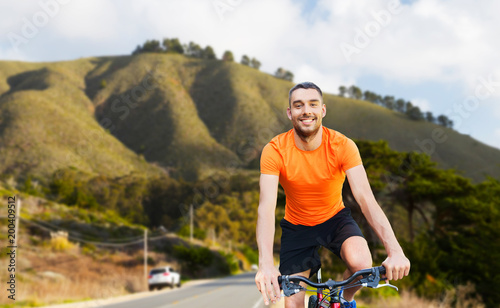 fitness, sport and healthy lifestyle concept - happy young man riding bicycle over big sur hills and road background in california