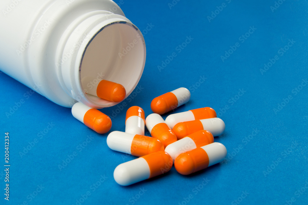 Orange white capsules (pills) were poured from a white bottle on a blue background. Medical background, template. Orange white capsules (pills) were poured from a white bottle on a blue background. Medical background, template.