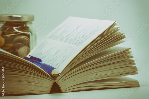 Opened book and a pen with blurred the glass jar of coin on dark background