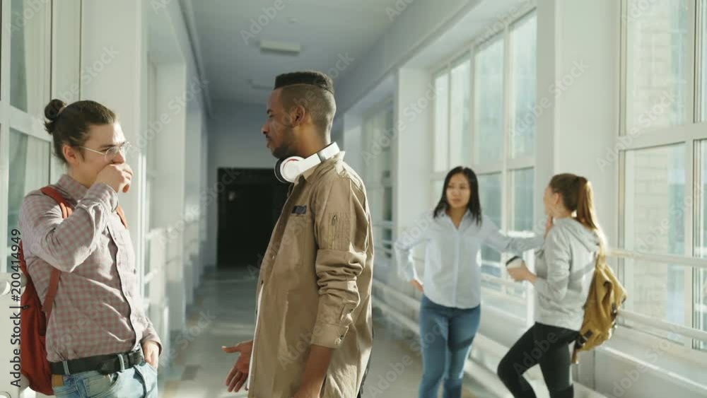 Multi-ethnic group of students talking in corridor of university having break. On front view guys are chatting to each other on back view girls are standing Multi-ethnic group of students talking in corridor of university having break. On front view guys are chatting to each other on back view girls are standing